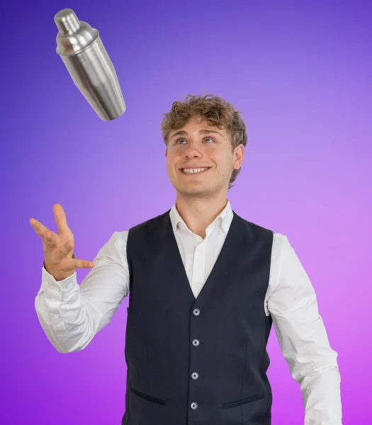 Junger Mann in weißem Hemd und dunkler Weste wirft einen Cocktailshaker in die Luft und schaut diesen an, vor violettem Hintergrund