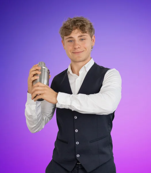 Vor einem violett-blauen Hintergrund steht ein junger Mann. Er lächelt, trägt ein helles Hemd und eine dunkle Weste. In beiden Händen hält er einen Cocktailshaker.