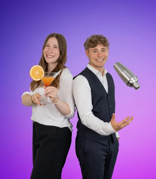Vor einem lila Hintergrund stehen zwei junge Personen. Links hält eine Frau ein Cocktailglas mit einer Orangenscheibe, während rechts ein Mann einen Cocktailshaker in die Luft wirft. Beide lächeln und tragen helle Oberteile; der Mann trägt zusätzlich eine dunkle Weste.