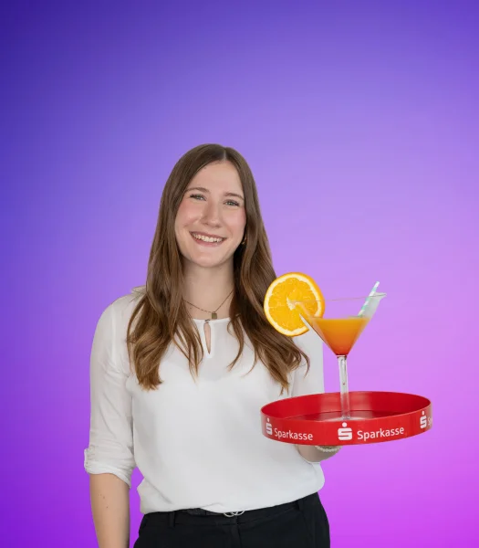 Eine junge Frau hält vor violettem Hintergrund ein Tablett mit einem Cocktailglas, in dem eine Orangenscheibe steckt, in der Hand.