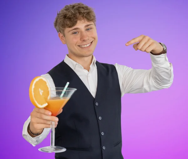 Lächelnder junger Mann im weißen Hemd und dunkler Weste hält ein Cocktailglas mit Orangenscheibe und zeigt mit dem Finger auf sich; violetter Hintergrund.