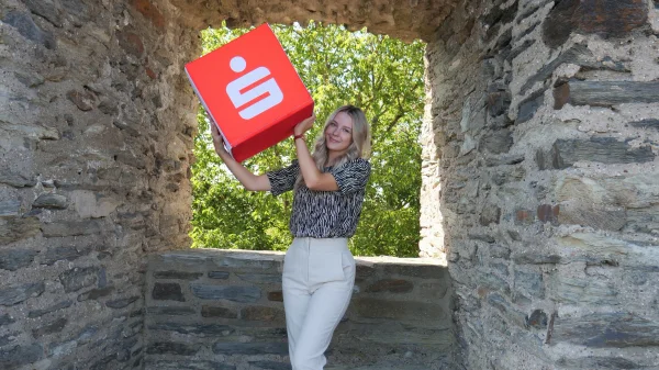 Eine Person steht in einem steinernen Torbogen und hält eine große rote Sparkassen-Box mit weißem Logo über der Schulter. Im Hintergrund sind grüne Bäume bei Tageslicht zu sehen.