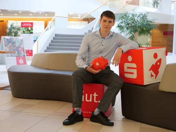 Ein junger Mitarbeiter sitzt im Foyer der Kreissparkasse auf einem roten Sitzwürfel und hält ein rotes Sparschwein in der Hand.